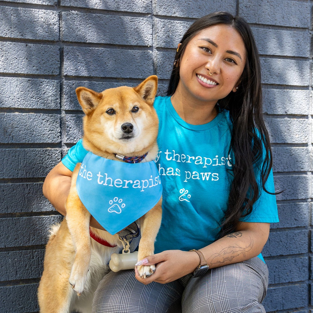 Woman in a blue 'therapist that paws' shirt with a Shiba Inu dog against a gray brick wall.